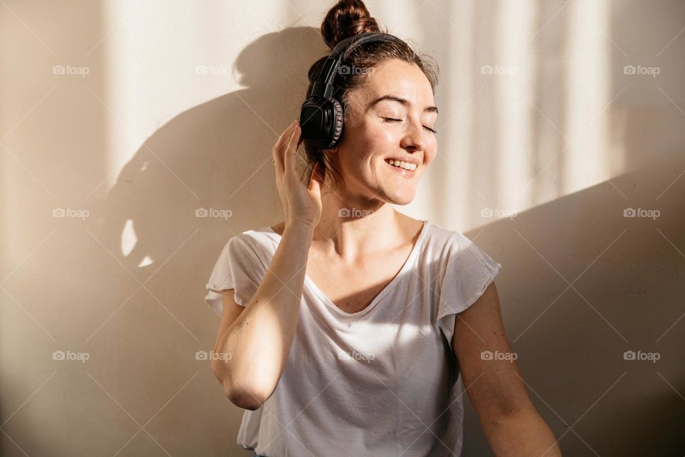 Young millennial woman, smiling and listening to music at home, at sunset time with a beautiful light on her.