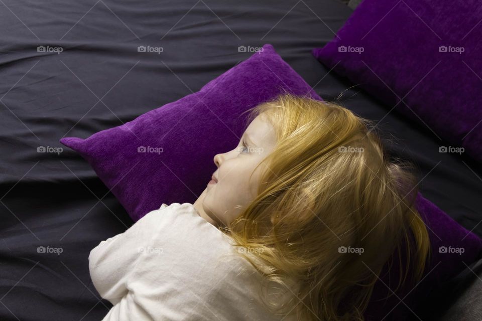 A little girl with red hair lies on a purple pillow.