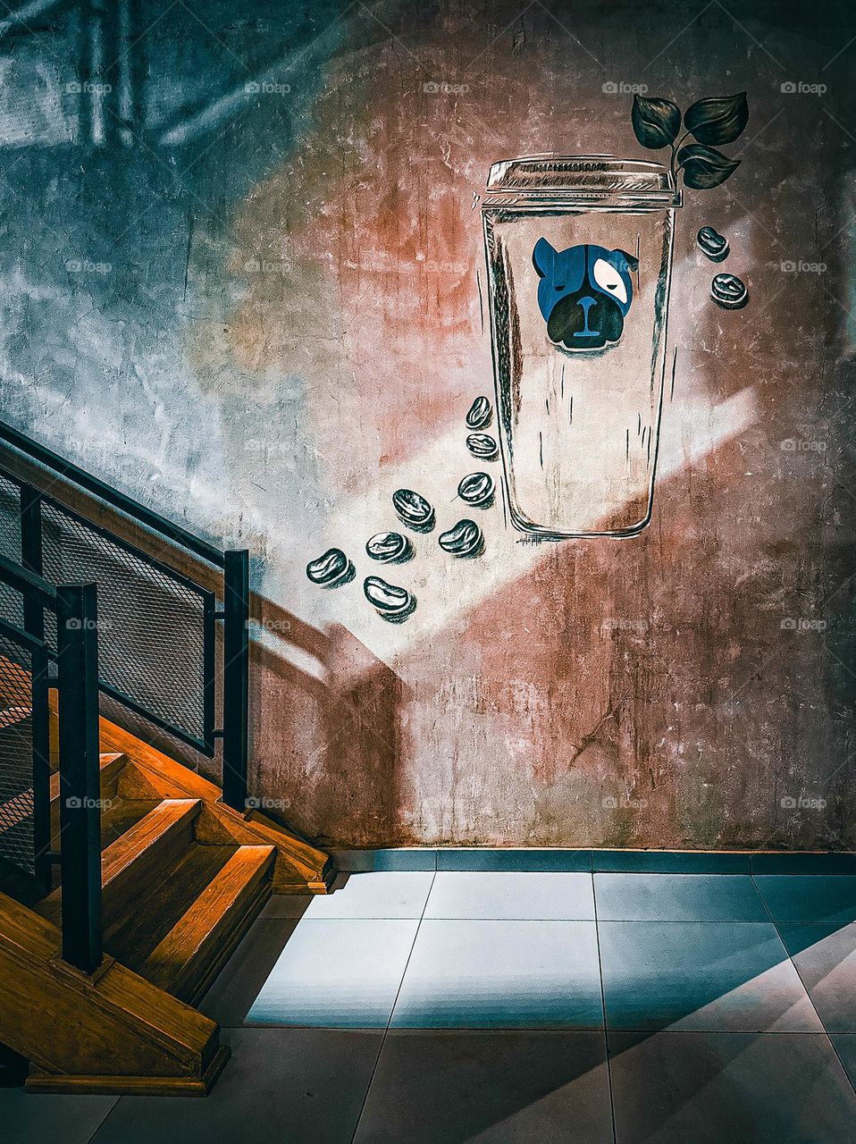 The photo captures a stairway corner illuminated by a dramatic interplay of light and shadow. On the adjacent wall, a painted coffee cup with a dog motif and scattered coffee beans adds an element of whimsy. 