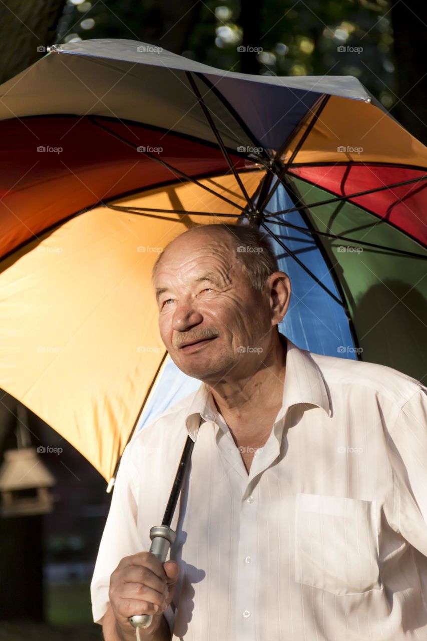 An elderly man of retirement age with wrinkles on his face stands under a multi-colored umbrella and smiles.