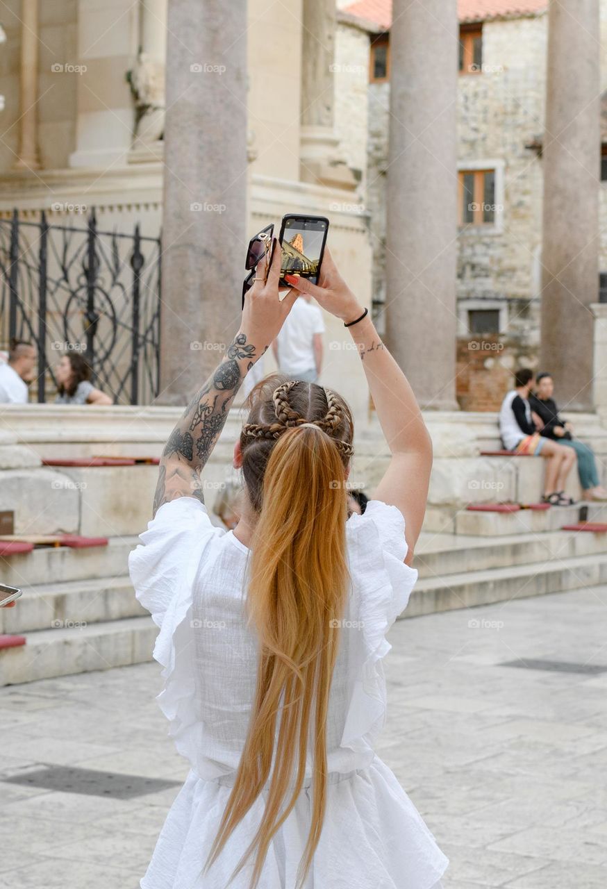 Rear view of woman with tattoo on her arm, taking photos of building in city