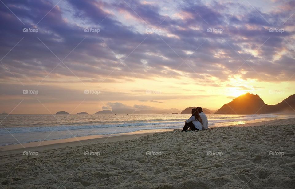 “The two sit alone on the beach and watch the sunset.
They look out over the water, the sun setting behind the hills to their right."