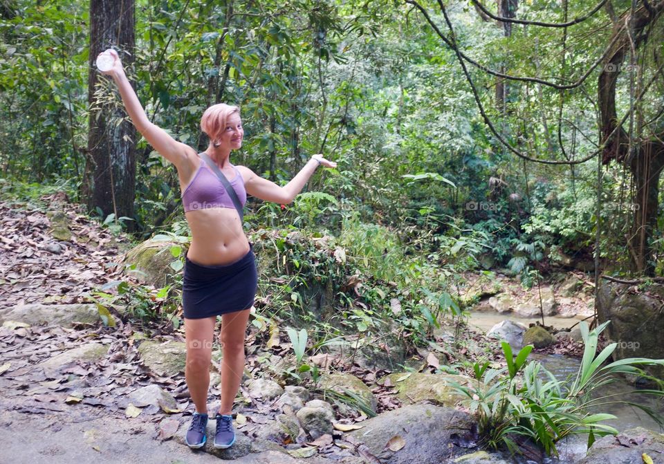 Woman wearing a black  skirt and a purple sports bra in the jungle with her arms in the air