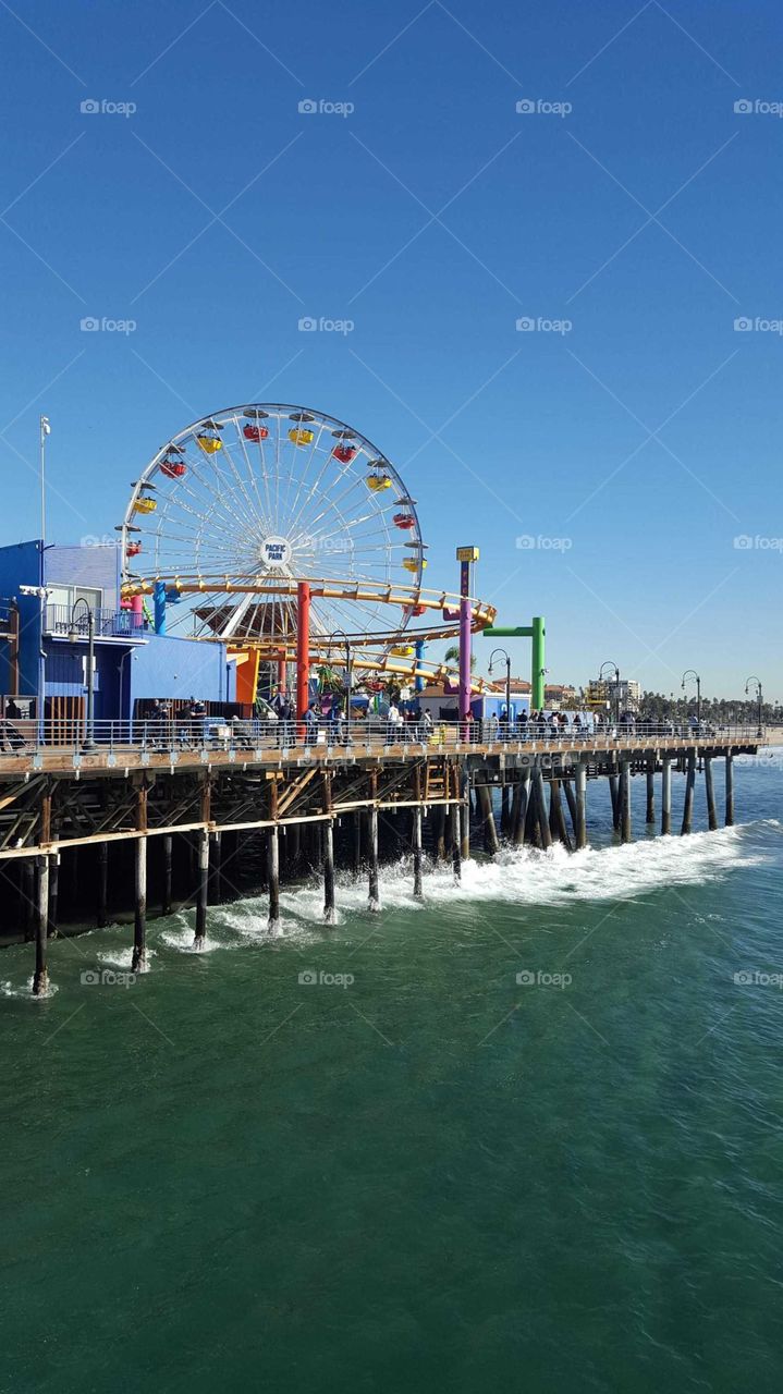 In this photo, the Santa Monica Pier in Santa Monica, CA, is on display. The beautifully colored ferris wheel, along  numerous rides and buildings, pop against the serene blue sky. The ocean waves clash against the brown, wooden pier.