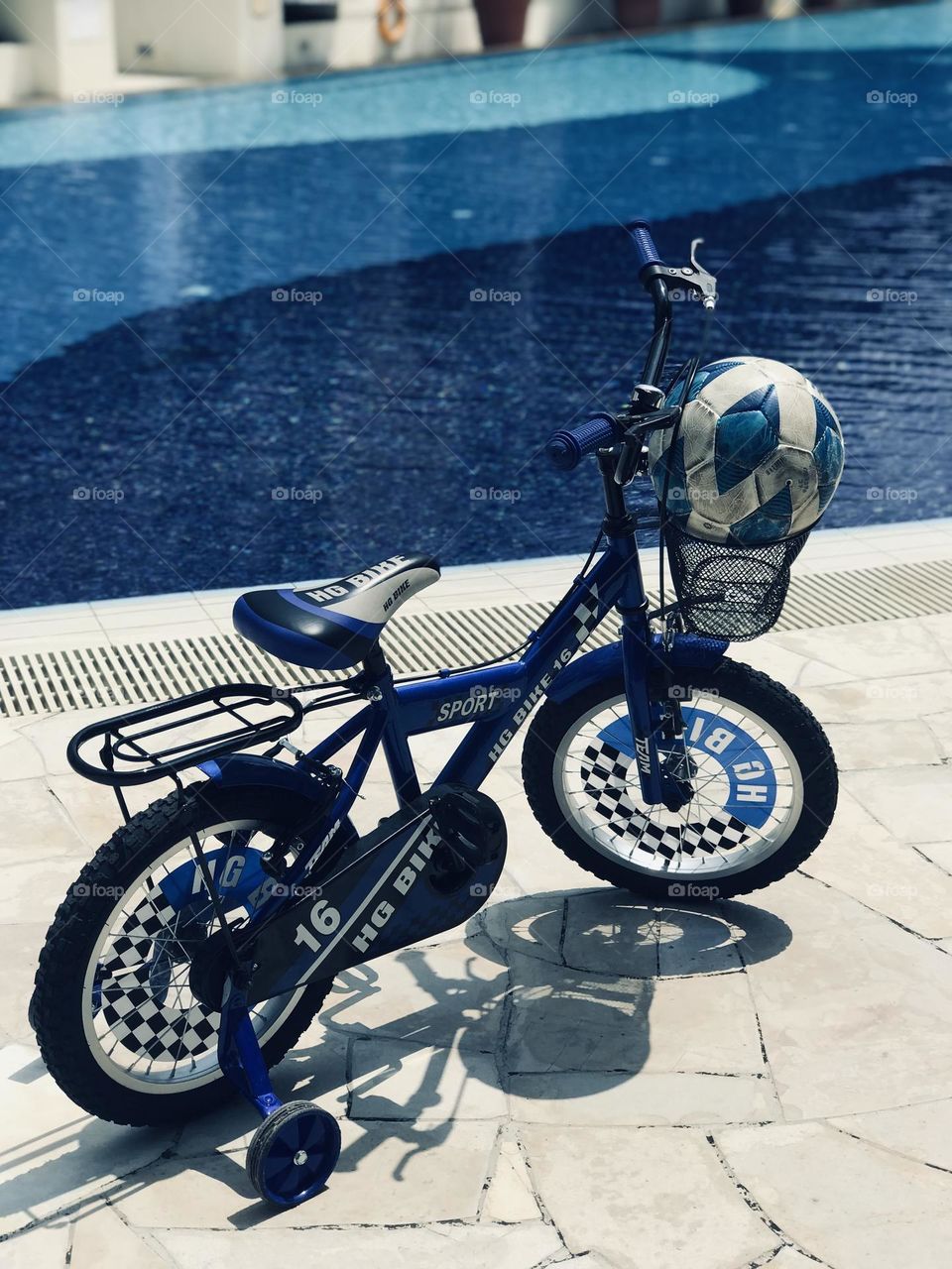 A blue kids bicycle with blue football in cycle basket stands near blue colour swimming pool.