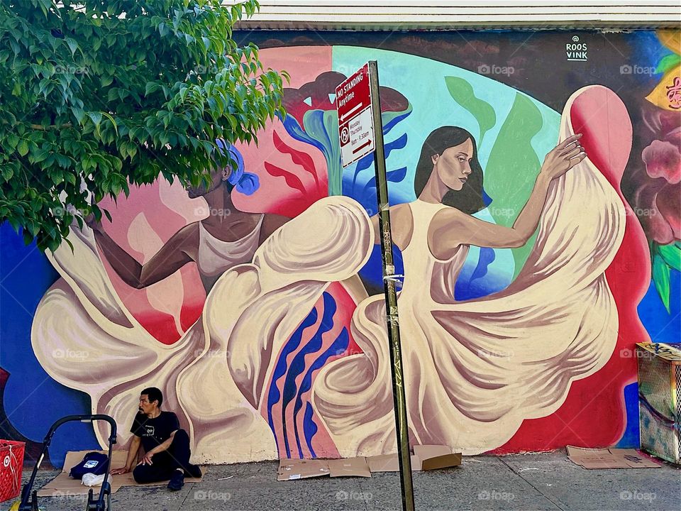 A man sits in front of the larger than life art mural on “Jefferson St” off “Broadway” in “Bushwick”, Brooklyn celebrating the Latin culture featuring pirouetting dancers in flowing white gowns. 2024. Hypnotic Productions