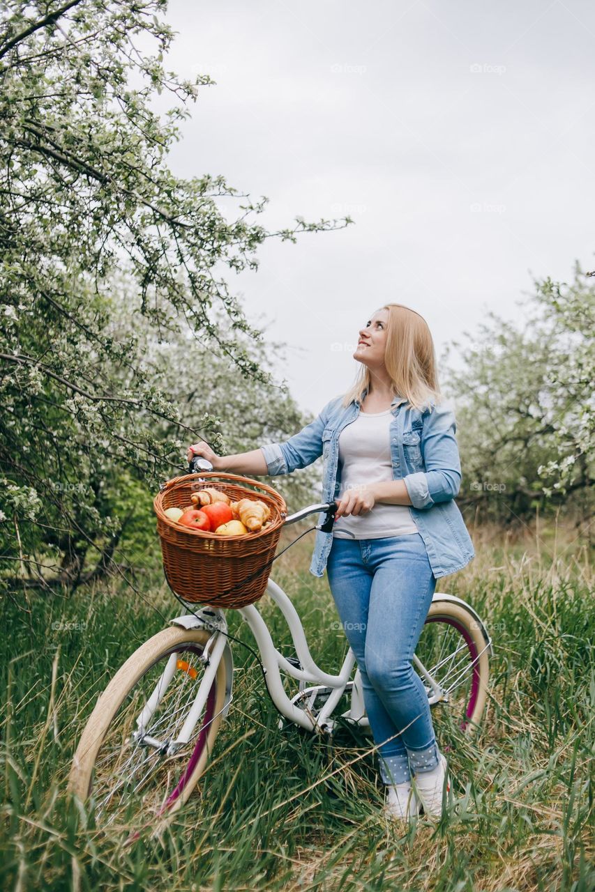 Blonde girl holding bicycle and looking to apple tree