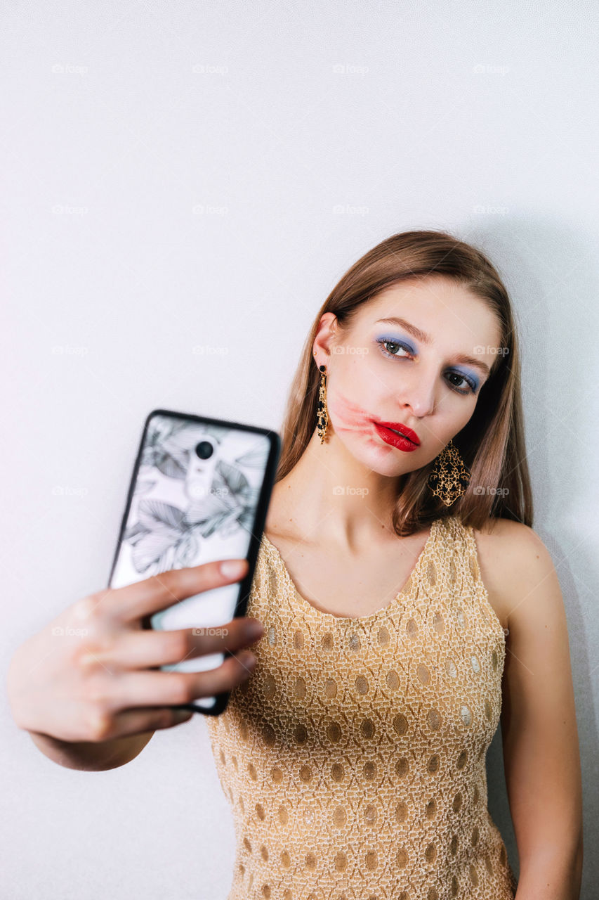 Portrait of a pretty blonde girl with smeared lipstick taking a selfie. She is holding a smartphone in one hand and looks at its camera. She is wearing long earrings (pendants). Her make-up is smeared but she looks confident.