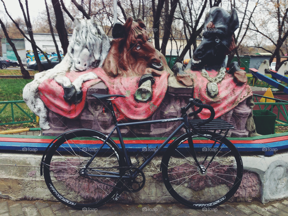 Grayish-black brakeless fixie bicycle standing near sculpture with three horses heads and a fence