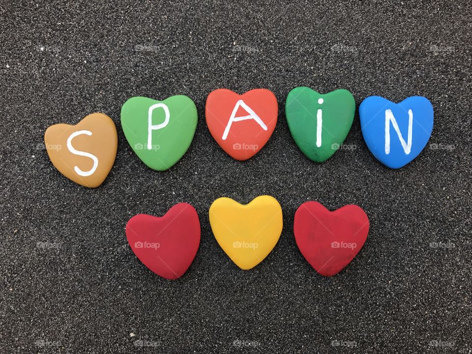 Spain, country name with colored heart stones over black volcanic sand