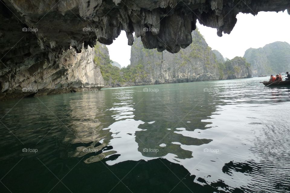 explore cave, on the ceiling of the jagged rock. dark gray color, lack of light, dark and cold. Outside, there is a boat rowing to carry tourists to visit. The mountains are reflected in the light blue water