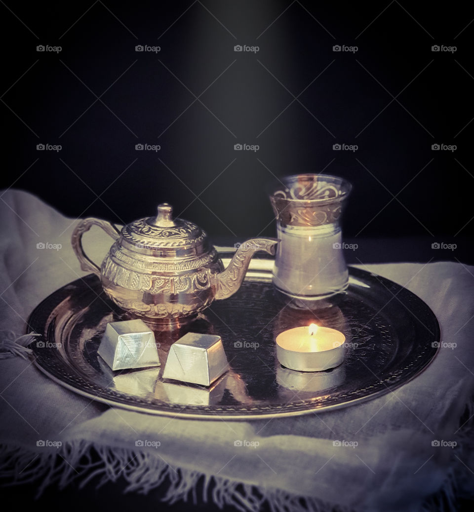 Dark oriental still life.  Silver ornamental tray with a small silver teapot and a glass of tea with milk, sweets and a burning candle on gray beautiful textile.  Tasty snack.  Food photo, square orientation