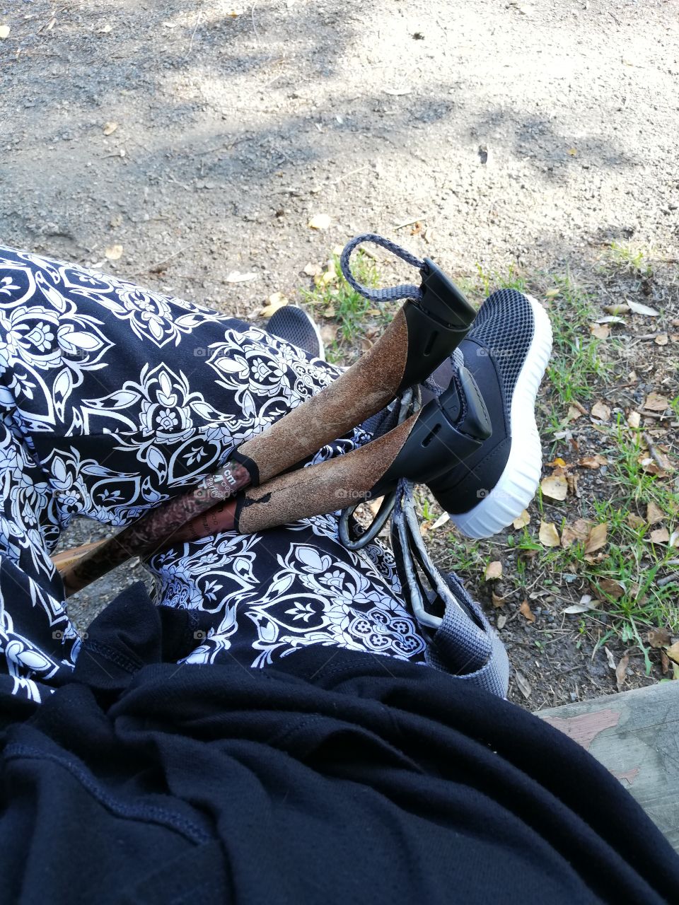 A person is sitting on the bench and enjoying the sunshine. The foot in the patterned pants is on another. The nordic walking poles are resting between the legs, the seam of the black cloth turned up.