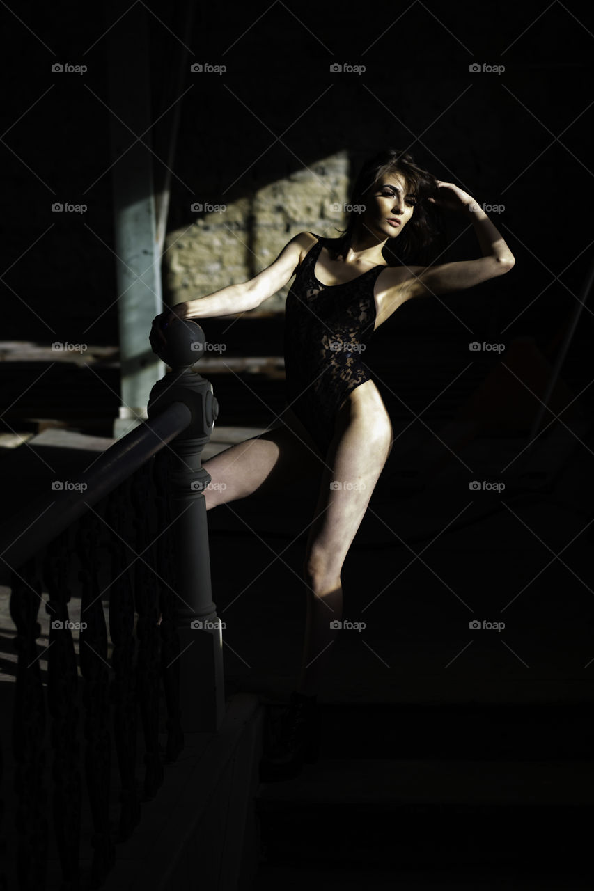 Model in lingerie standing on the stairs. She is holding on to the hand rail. She is lit by natural light and is partly in shadow with great contrast