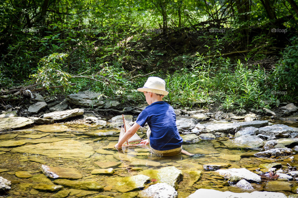 Boy playing with a toy sailboat in a creek during the summer