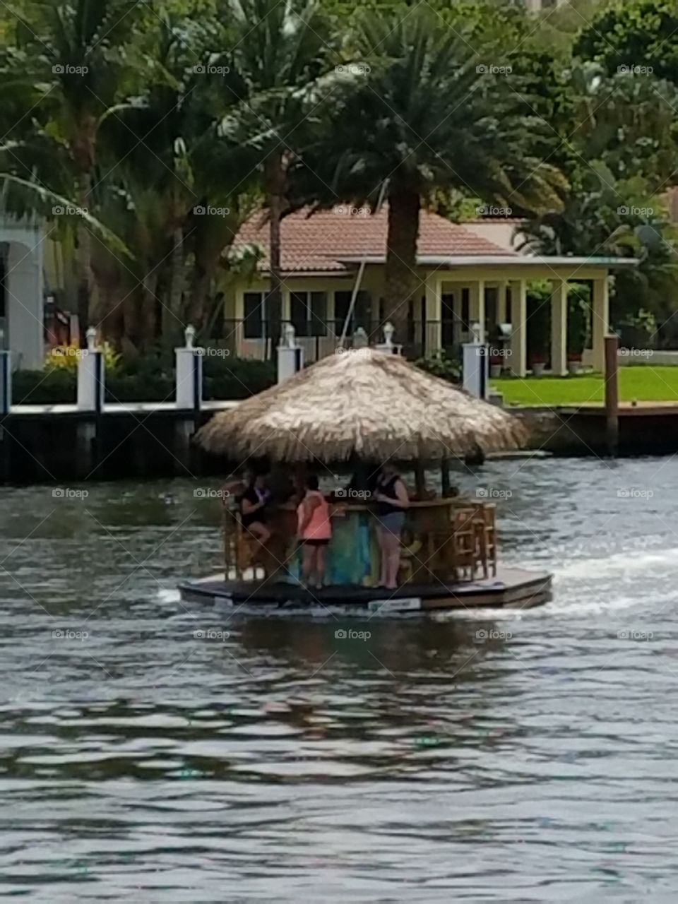 People sitting on stools at bar, drinking. Boat is round, thatched roof & engine running down river. Happy people drinking having fun.