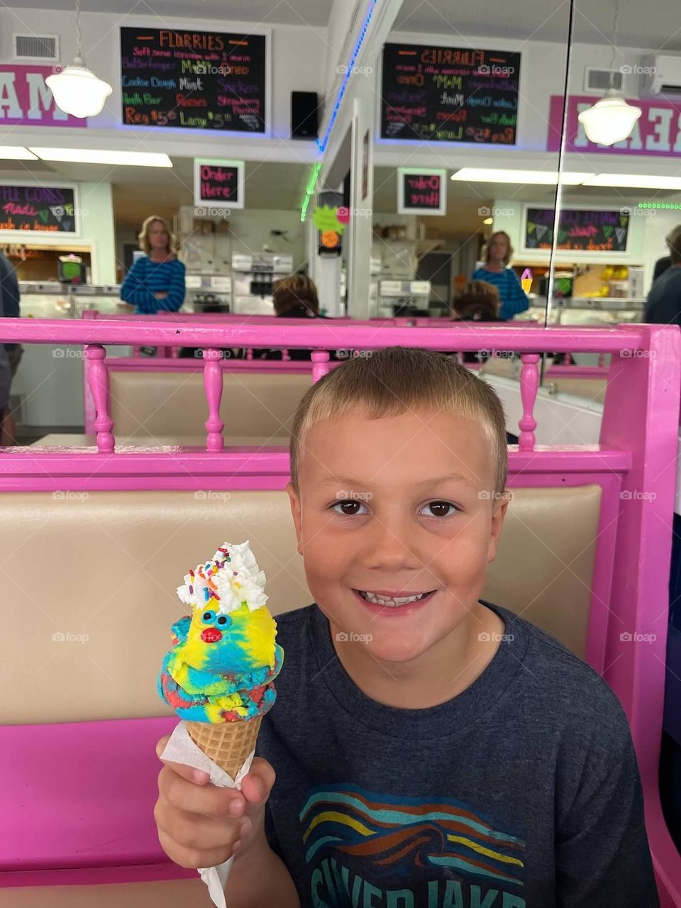 A young boy smiles as he holds his blue, yellow, and red ice cream topped with whipped cream and sprinkles in a freshly made warm waffle cone. A pink booth evokes feelings of joy. The sweet creation looks like a duck with its candied eyes and bill!