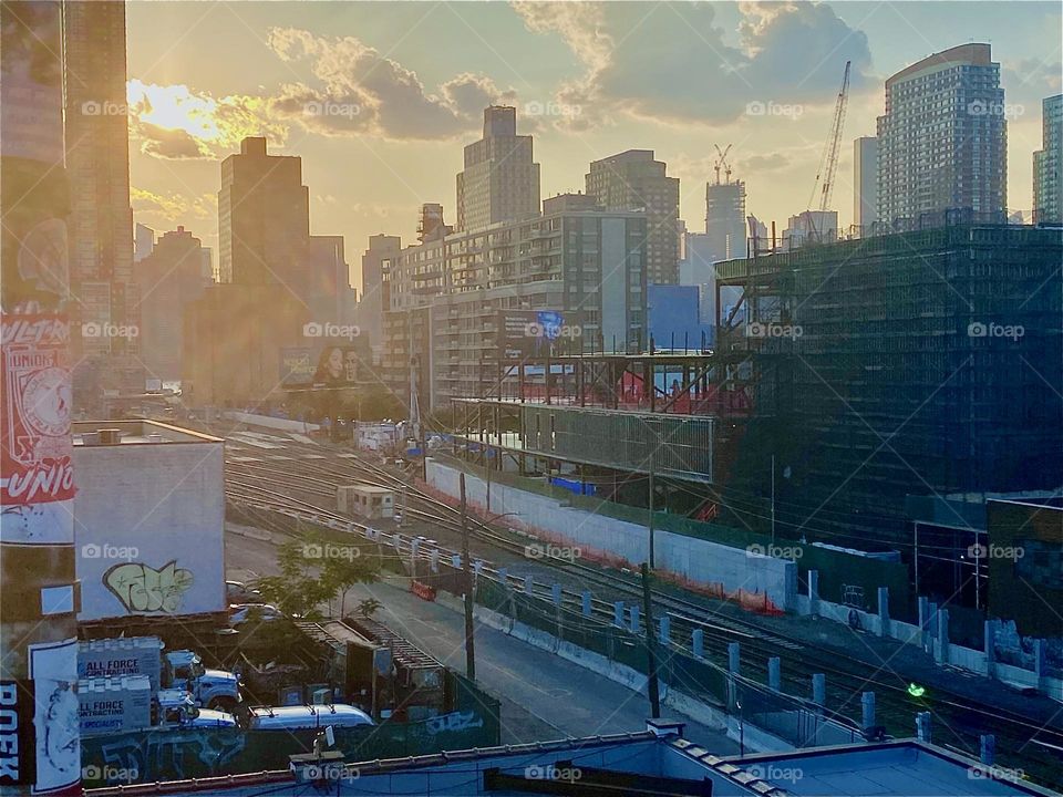 This is a view from the „Pulaski Bridge“ LIC, Queens across the train tracks onto „Greenpoint“, Brooklyn and in the far distance „Manhattan“ during the golden hour of sunset on a slightly hazy day. Perhaps it’s smog. 2023. Hypnotic Productions