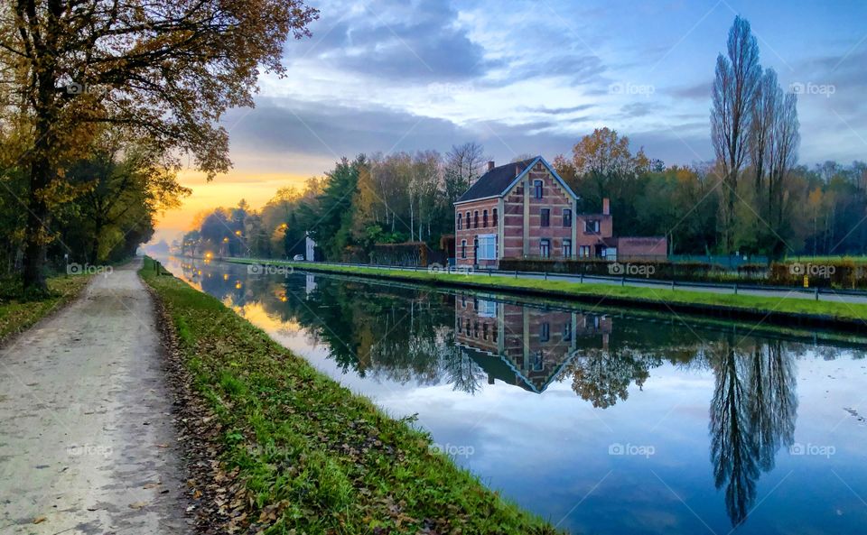 Cosy house reflected in the water of the river or canal that flows through a forest area, all this under a sunset or sunrise sky