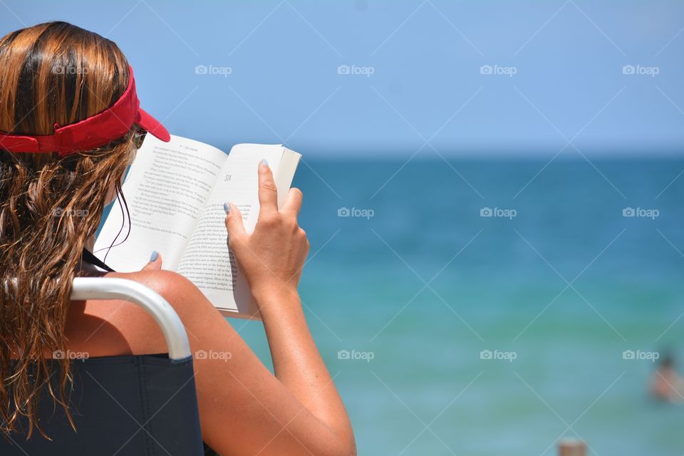 Relaxing with a good summer read, a female reading a book at the beach with tropical ocean water in background.