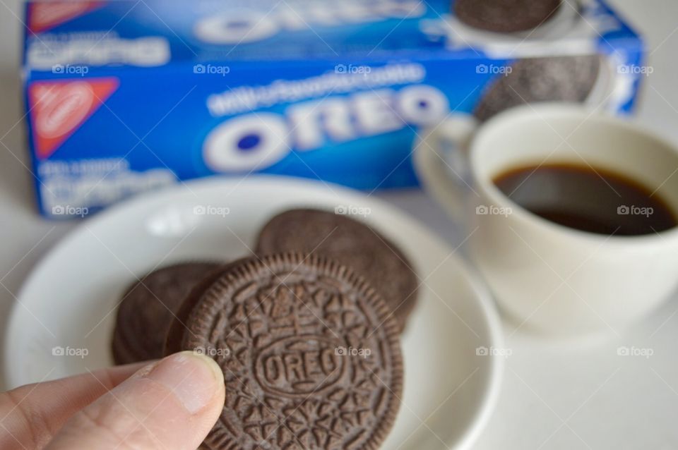 A persons fingers holding an Oreo cookie and a cup of coffee in the background