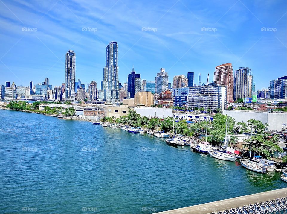 A panoramic view from the “Pulaski Bridge” at “Newtown Creek” overlooking the “East River” onto “LIC, Queens” & “Manhattan” from further away, beautiful boats & sailboats lining the shore with its lush green vegetation. 2022. Hypnotic Productions
