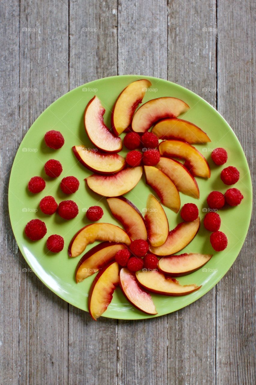 Flat lay of a decorative arrangement of fresh nectarine slices and raspberries on a green plate on a rustic, light, wooden background 