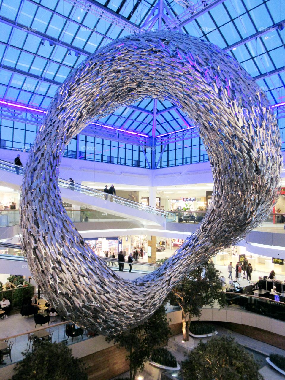 Shopping centre with glass roof and silver fish making it look like they are swimming in a shoal with shops and escalators in background