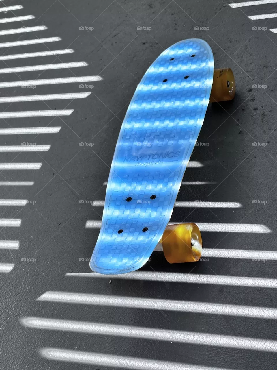 Kid’s blue poly Kryptonite short board skateboard with urethane wheels in shadow of fence with shadow stripes and light