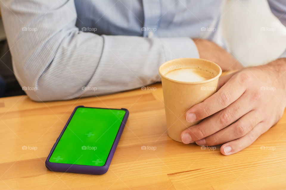 Close-up of male hands in a cafe hold a cup of coffee in his hand. Morning routine lies next to a phone with a green mocap screen. Concept: lifestal life businessman routine morning