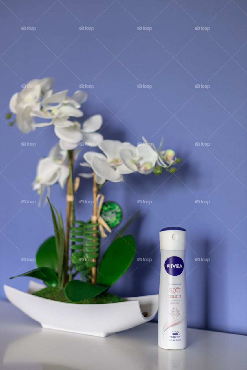 One bottle of Nivea brand anti transpirant with a potted orchid flower stands on a white chest of drawers in a bedroom against a lilac blue wall, close-up side view.