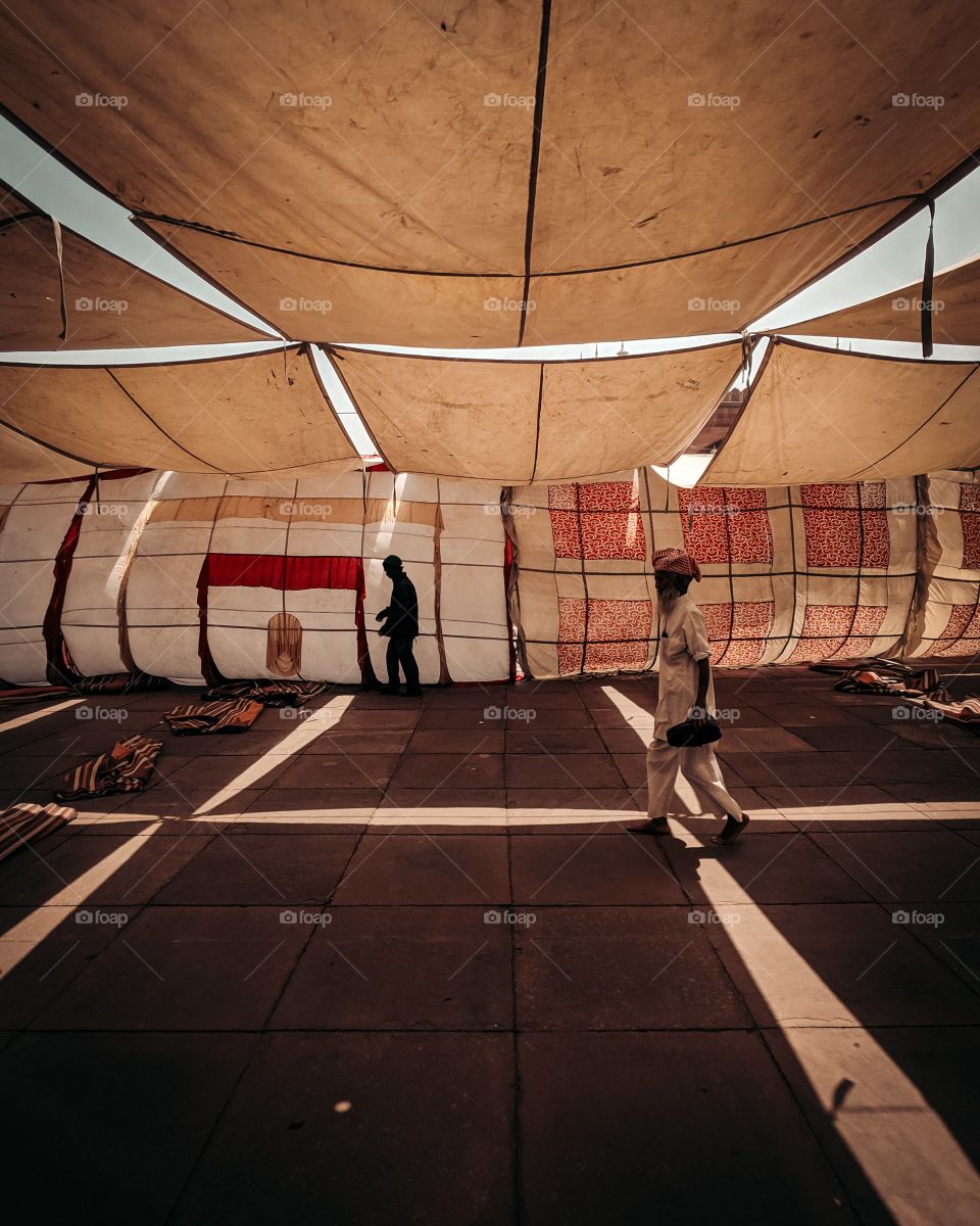 Two people walk under a large tent connected by different pieces of fabric creating a canopy against the scorching sun.