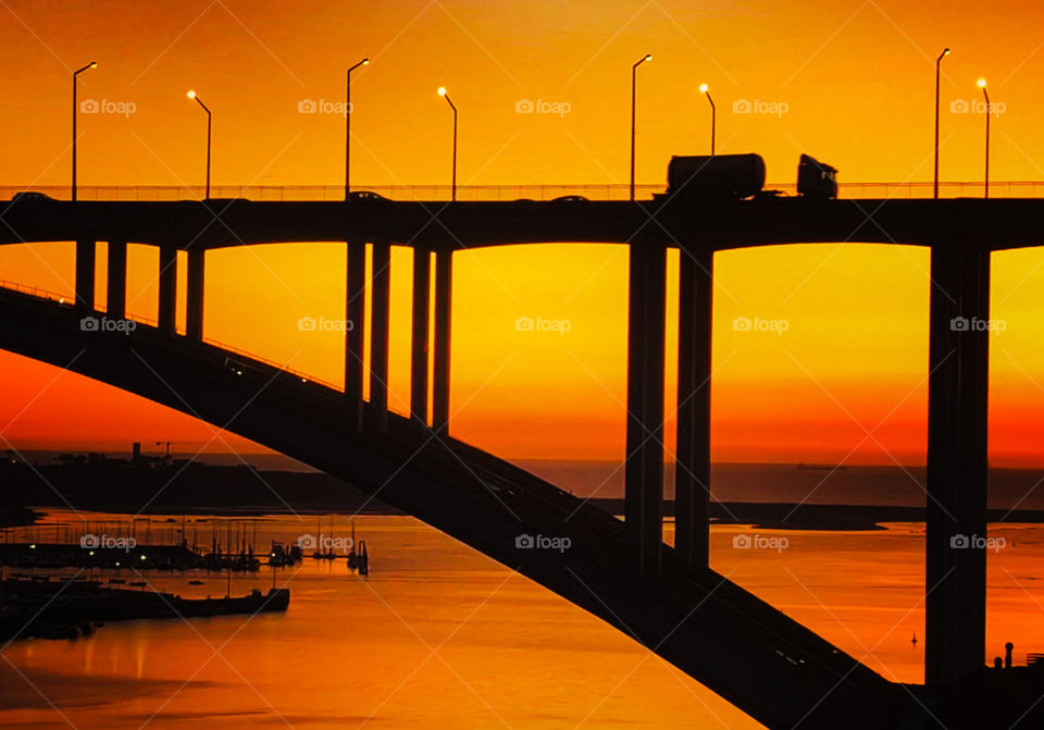 A deep orange sunset silhouettes the traffic crossing Ponte da Arrábida in front of the mouth of Rio Douro, Porto, Portugal 