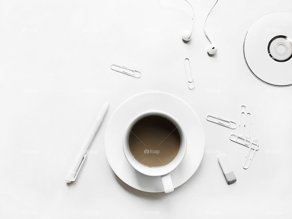 White Cup of coffee with milk on the white saucer, white pen, white clips, white cd, white headphones and white rubber in white table 
