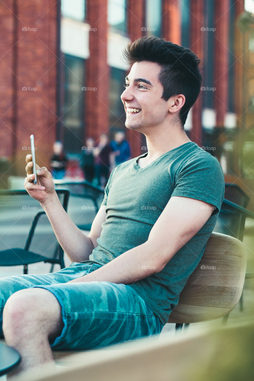 Young man having fun with smartphone, reading funny texts, sitting in center of town