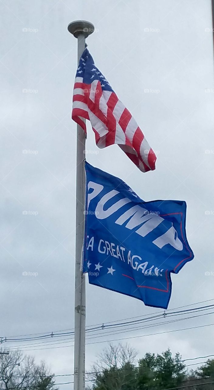 Photo of our flagpole flying 🇺🇸 USA Flag & President Trumps MAGA flag flying against a gray cloudy sky.
