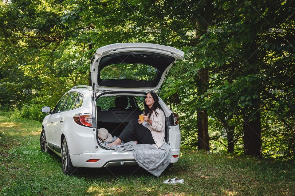 Young woman drinking coffee while sitting in trunk of a car parked in woods