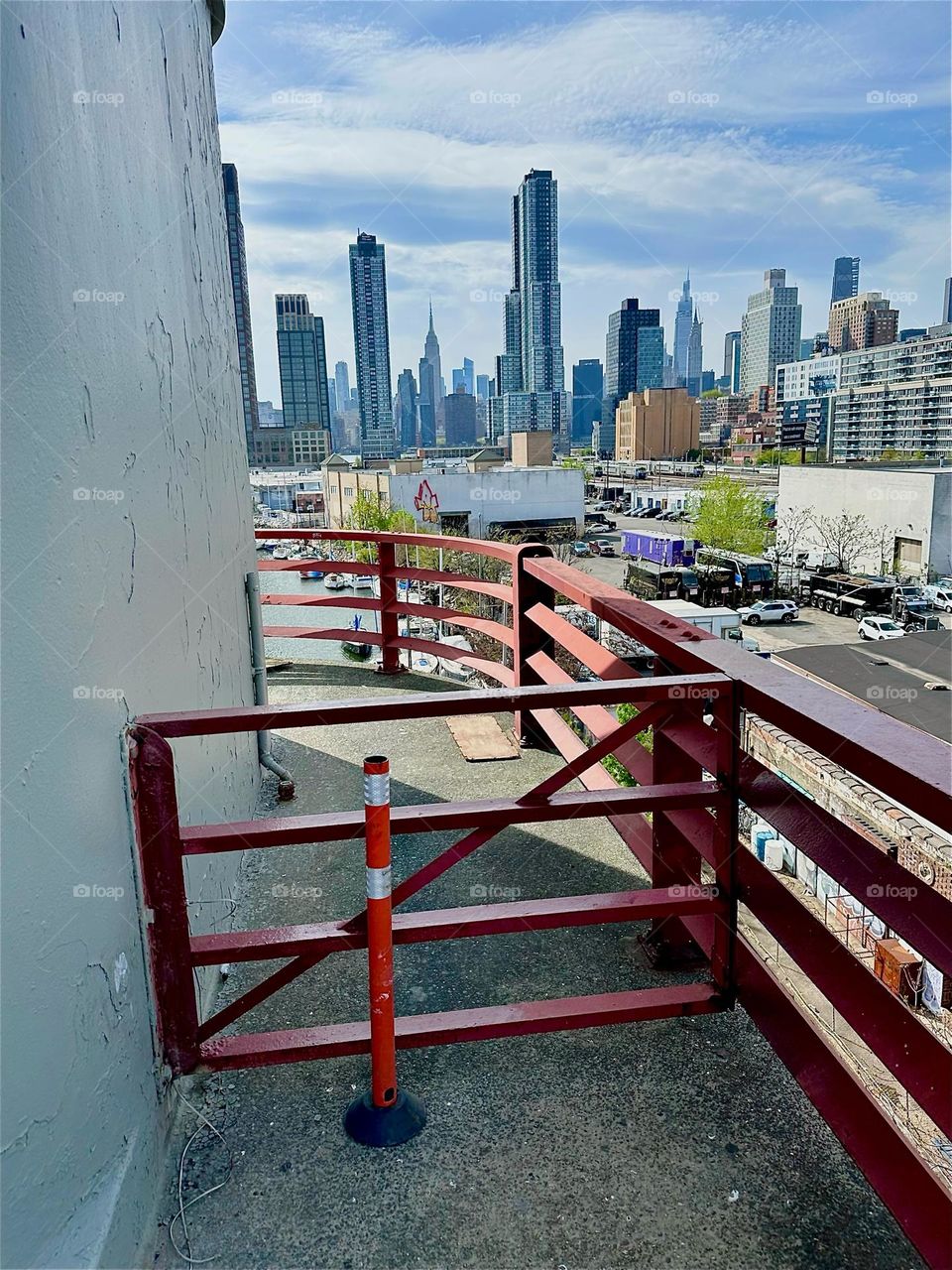 The orange red of the “Bauhaus” curved metal railings of the “Pulaski Bridge” that connects LIC to “Greenpoint”, Bklyn is juxtaposed to its complementary color blue in the LIC and “Manhattan” high rises and the sky above. 2025. Hypnotic Productions