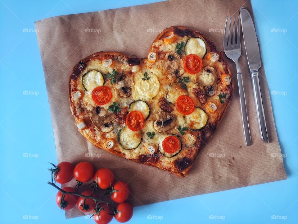 Delicious vegetarian pizza in the shape of a heart for Valentine's Day with tomatoes, vegetables and cheese on a blue background.