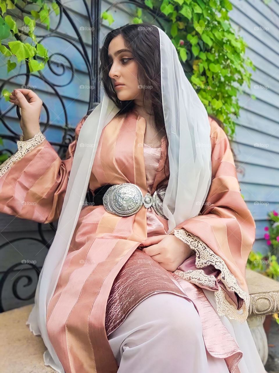 Close-up of a young woman wearing traditional pink and white clothing, sitting in front of a background of green plants and blue walls. The outfit has delicate embroidered details and a large silver belt