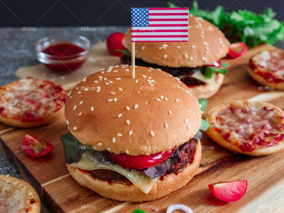 One large and juicy burger with an American paper flag on a toothpick rests on a wooden board on a stone background,close-up side view.