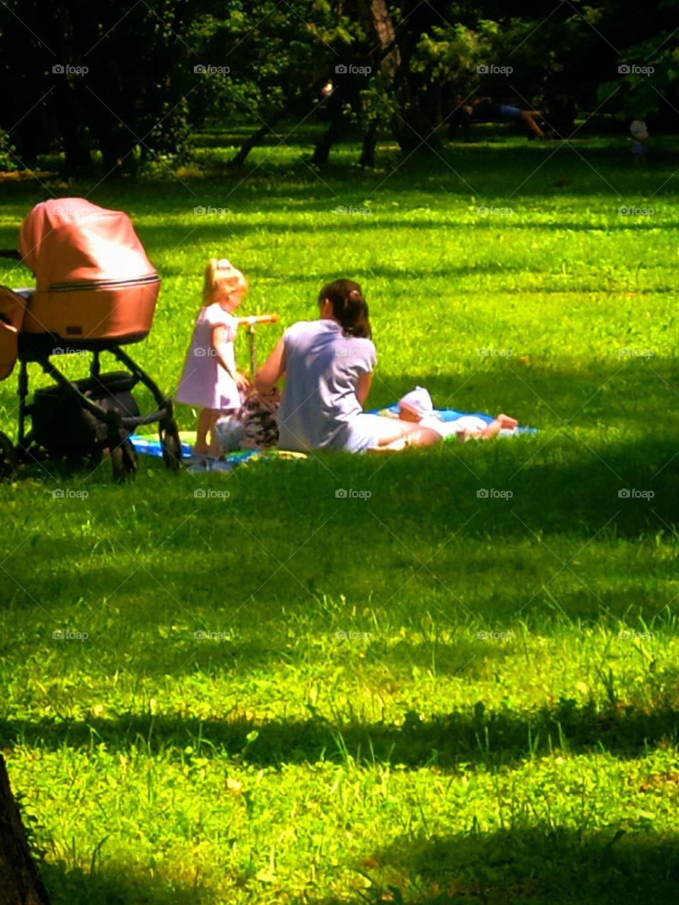 Summer in the city. Hot day. Green lawn in the park. On the green grass, in the shade of trees, a young woman sits, next to whom lies a baby and stand a little girl with a scooter. A baby stroller is standing next to a woman