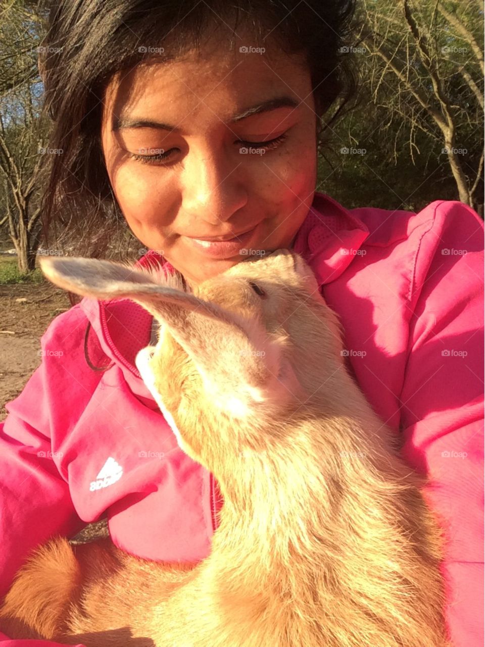 Close up of a young girl with dimples while she cradles a baby goat. Goat kid has a golden color & is lovingly looking up at the girl. The girl is looking down at the kid with a smile on her face. Set outdoors.
