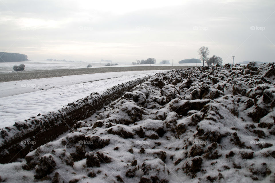Acker Boden gepflügt am Wegrand vor Landschaft mit Schnee Wolken Bäumen und Asphalt Weg.