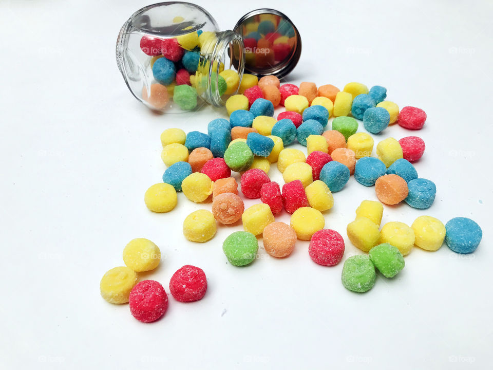 Sour chewy gummies in green, turquoise, red, yellow & orange flowing out of a small overturned glass jar. The up turned lid reflects the candies. The candies have little smily faces & an orange one in the foreground smiles at the viewer! ๐