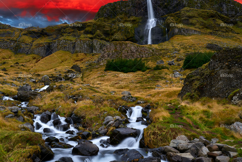 Idyllic stream flowing from the impressive Bjarnafoss waterfall in Snaefellsnes Peninsula Iceland on Autumn afternoon in October 2019. This is a composite image with red sky taken from a different photo with dramatic clouds at sunset over lake.