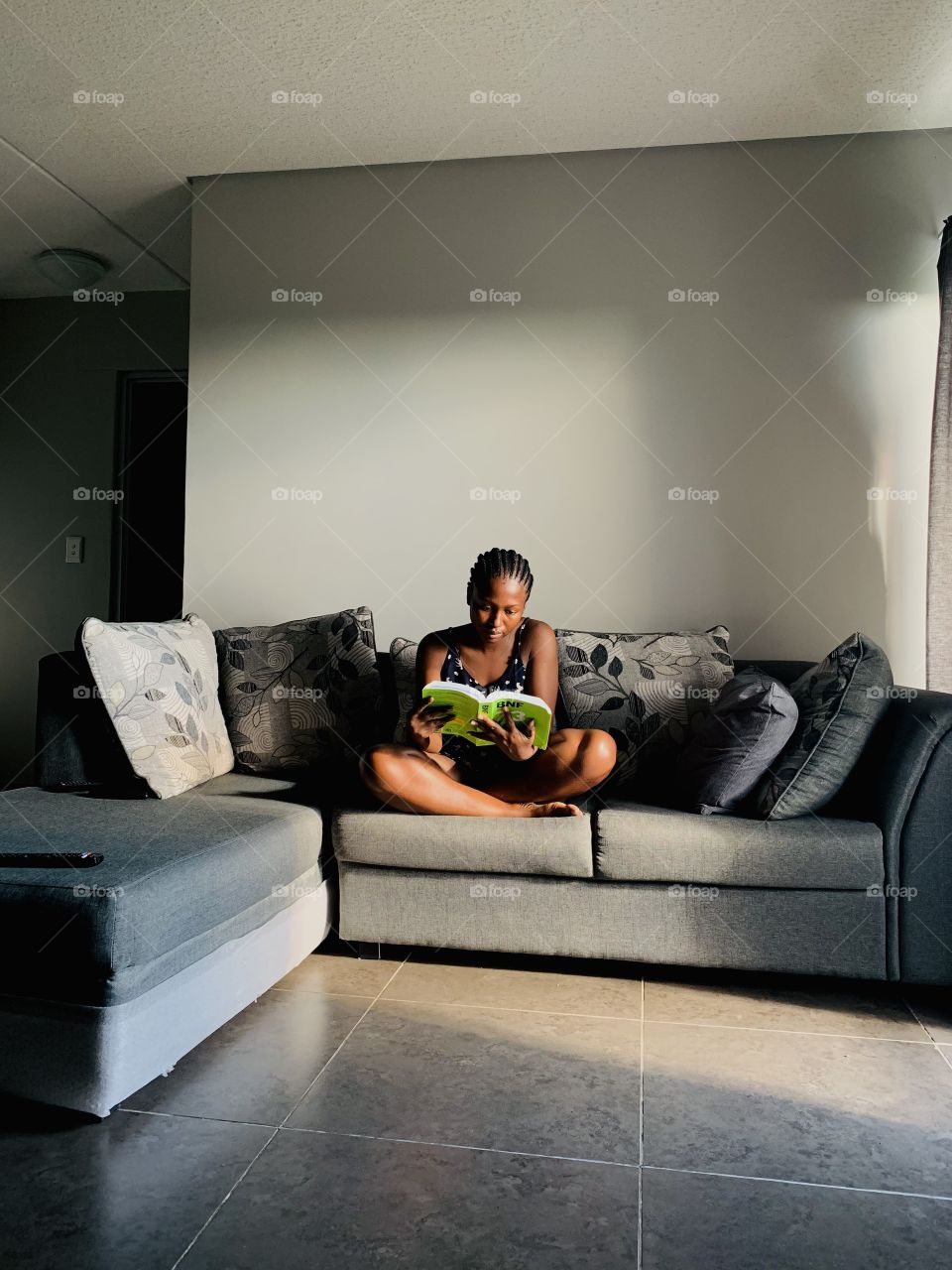 A black woman sitting on a grey couch with grey floral cushions, while reading her book.