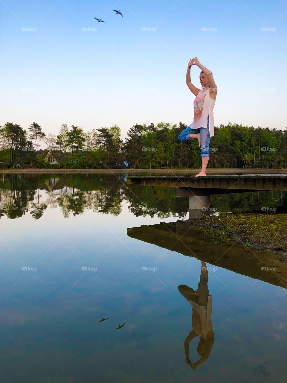 An attractive young woman doing an asana yoga pose, the tree with variation, vrksasana, for balance and stretching near the lake with reflection in the water.