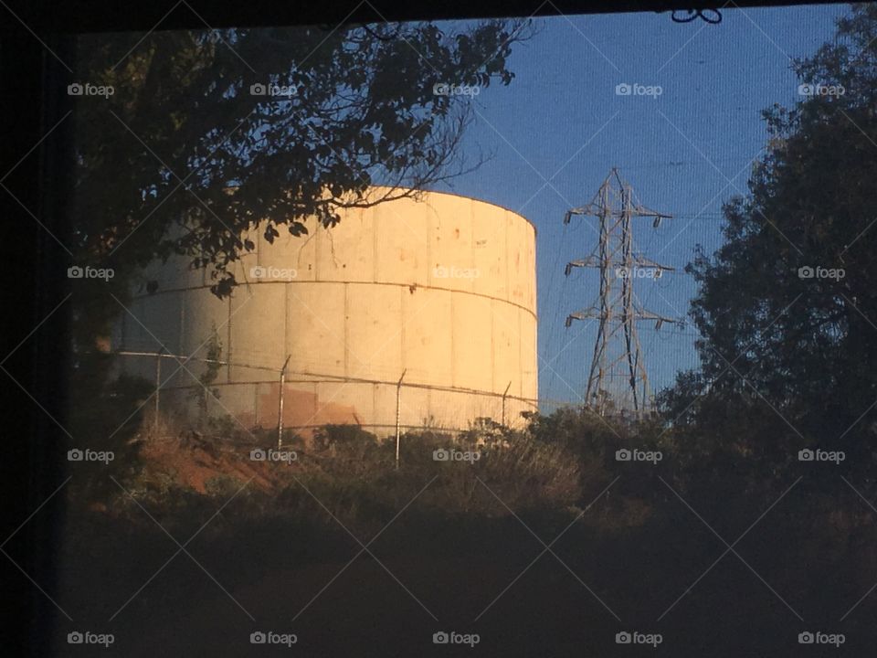 Rustic water tank is lighted by the setting sun. A tree and power line tower frame the scene on a California day 