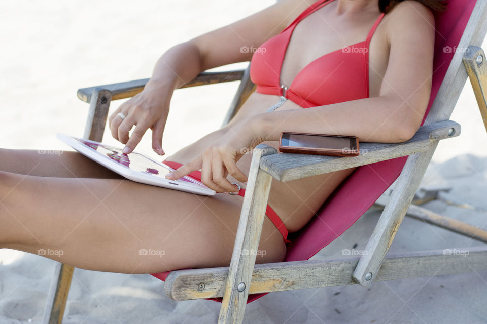 woman using tablet on beach. woman in red bikini using her white tablet sitting in deck chair on beach